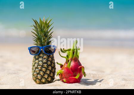 Ananas mûr attrayant en lunettes de soleil et fruit dragon rose sur la plage tropicale de sable contre l'eau de mer turquoise, Thaïlande. Vacances d'été concep Banque D'Images