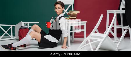 étudiant élégant en lunettes tenant une boisson dans une tasse en plastique près des chaises et des livres sur fond vert et bordeaux, bannière, image de stock Banque D'Images