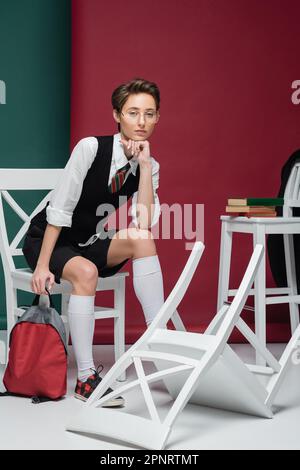 jeune étudiant en lunettes assis avec un sac à dos autour des chaises et des livres sur fond vert et marron, image de stock Banque D'Images