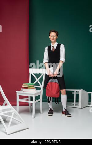 pleine longueur de jeune étudiant élégant en lunettes debout avec sac à dos autour des chaises et des livres sur fond vert et rose, image de stock Banque D'Images