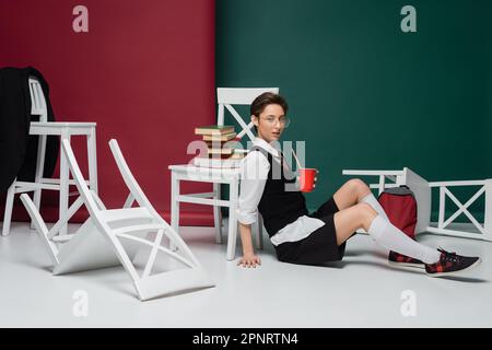 jeune étudiant élégant en lunettes tenant une boisson dans une tasse en plastique près des chaises et des livres sur fond vert et bordeaux, image de stock Banque D'Images