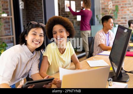 Portrait de deux collègues créatifs de sexe féminin, souriant à leur bureau, dans un bureau animé et décontracté Banque D'Images