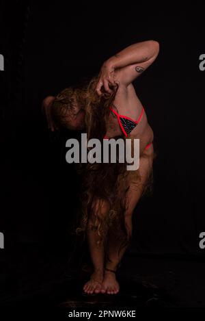 Jeune homme, acteur, portant bikini dans performance gorilla femme avec les cheveux partout dans son corps et expression de la douleur. isolé sur fond sombre. Banque D'Images