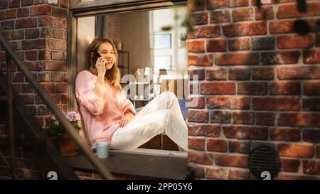 Belle jeune femme ayant un appel sur son smartphone pendant qu'elle est assise sur son rebord de fenêtre. Femme expressive parlant au téléphone, parlant. Banque D'Images