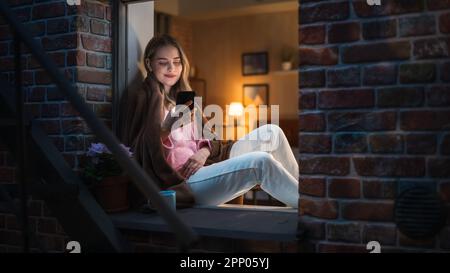 Belle femme blanche utilisant un smartphone tout en étant assise sur un rebord de fenêtre ouvert. Belle fille avec style de vie créatif sain se détend à la maison Banque D'Images