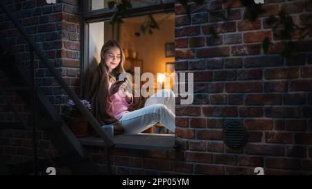 Belle femme blanche utilisant un smartphone tout en étant assise sur un rebord de fenêtre ouvert. Belle fille avec style de vie créatif sain se détend à la maison Banque D'Images