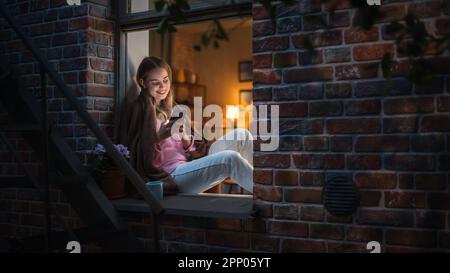 Belle femme blanche utilisant un smartphone tout en étant assise sur un rebord de fenêtre ouvert. Belle fille avec style de vie créatif sain se détend à la maison Banque D'Images