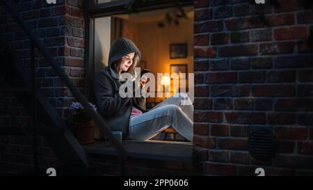 Femme blanche portant un sweat à capuche à l'aide de son smartphone tout en étant assise sur le rebord de la fenêtre la nuit. Jeu de femmes jouant à des jeux de téléphone mobile addictifs à la place Banque D'Images