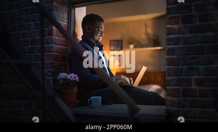 Jeune beau Black Man travaillant à la maison sur ordinateur portable dans la fenêtre d'un confortable appartement loft élégant. Homme souriant, vérifier les médias sociaux Banque D'Images