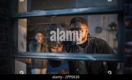 Jeune homme noir expliquant une équation mathématique sur une fenêtre à la maison à un ami féminin en utilisant des marqueurs de verre effaçable. Deux étudiants multiethniques Banque D'Images