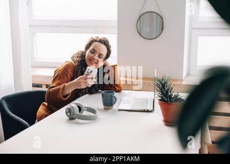 Jeune heureuse souriante jolie femme assise sur une chaise tenant un smartphone à l'aide de la technologie mobile de téléphone portable regardant un téléphone mobile tout en étant à distance Banque D'Images