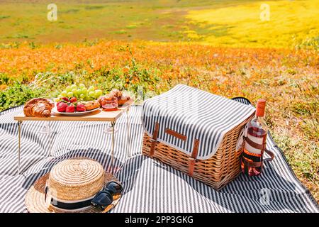 Composition de pique-nique d'été, panier en osier, chapeau de paille, vin et petite table avec fruits et croissants sur prairie de fleurs Banque D'Images