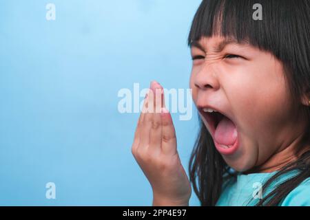 Petite fille asiatique couvrant sa bouche pour sentir le mauvais souffle. Enfant fille vérifiant la respiration avec ses mains. Problèmes de santé bucco-dentaire ou concept de soins dentaires. Banque D'Images