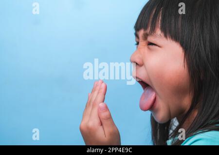 Petite fille asiatique couvrant sa bouche pour sentir le mauvais souffle. Enfant fille vérifiant la respiration avec ses mains. Problèmes de santé bucco-dentaire ou concept de soins dentaires. Banque D'Images