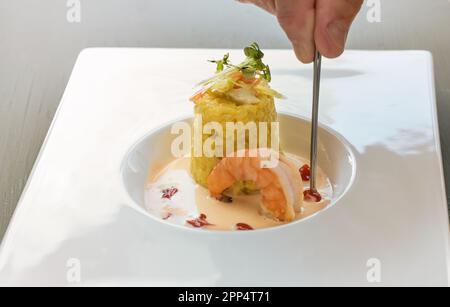 À la main avec une pince à épiler, un plat gastronomique de risotto au safran avec une garniture aux herbes, une grande sauce aux crevettes et aux crustacés dans une assiette blanche moderne, copie Banque D'Images