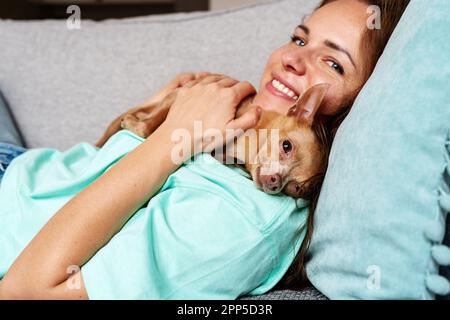 Une femme heureuse et son chien de mer Toy Terrier allongé sur un canapé à la maison. Banque D'Images