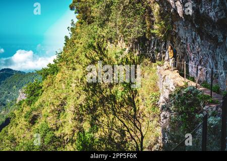 Description: Femme touristique bénéficiant d'une vue depuis le dessous d'un grand mur de roche le long du canal d'eau à la falaise abrupte à travers la forêt tropicale de Madère. Levada de Caldeir Banque D'Images