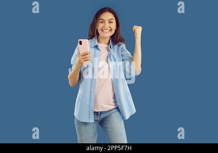 Une femme heureuse se réjouit sincèrement de la bonne nouvelle tout en regardant le téléphone mobile sur fond bleu. Banque D'Images
