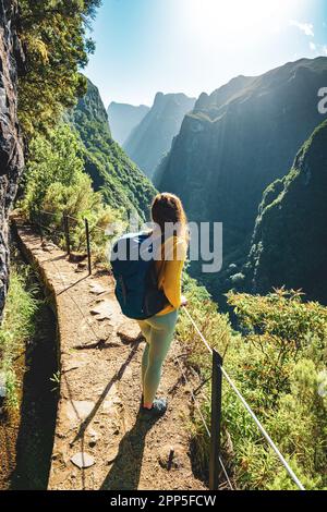 Description: Backpacker femme jouissant d'une vue panoramique depuis le dessous grand mur de roche le long du canal d'eau à la falaise raide à travers la forêt tropicale de Madère. Levada Banque D'Images