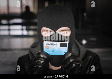 Criminel masqué qualifié utilisant une carte de crédit volée pour acheter des choses en ligne. Un voleur dans un masque noir tient une carte bancaire volée dans ses mains. Banque D'Images