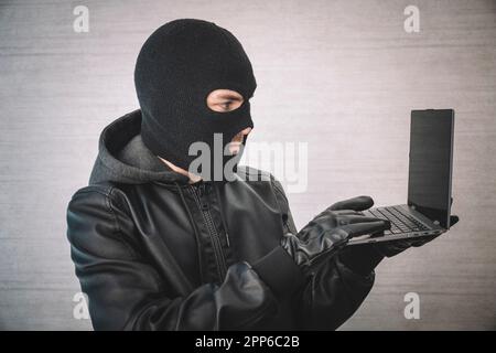 cybercriminalité, piratage réfléchi et criminalité technologique : pirate informatique dans une salle blanche avec ordinateur portable, pirate informatique dans un masque noir avec un ultrabook moderne, Banque D'Images