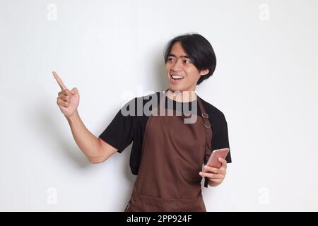 Portrait d'un barista asiatique attrayant en tablier marron tenant et pointant un téléphone mobile. Image isolée sur fond blanc Banque D'Images