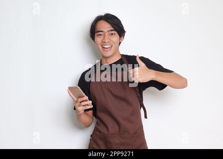 Portrait d'un joli barista asiatique en tablier marron montrant le pouce vers le haut tout en tenant un téléphone portable. Concept publicitaire. Imag. Isolée Banque D'Images