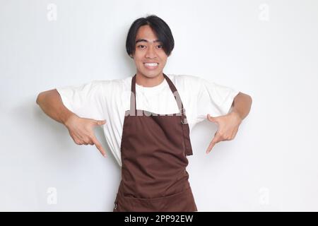 Portrait d'un barista asiatique attrayant en tablier marron montrant le produit, pointant vers quelque chose avec les mains. Concept publicitaire. Image isolée sur le coup Banque D'Images