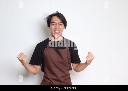 Portrait d'un barista asiatique en tablier marron qui élève son poing pour célébrer son succès. Image isolée sur fond blanc Banque D'Images