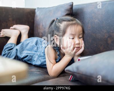 Belle jeune fille enfant s'allonger sur un canapé à la maison, regarder une tablette, fille asiatique Banque D'Images