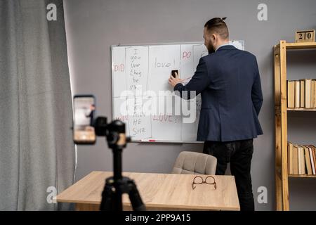 Professeur de sexe masculin debout près du tableau noir avec règles de grammaire anglaise et ayant une leçon en ligne. Tuteur millénaire donnant des cours de langue étrangère sur vidéo Banque D'Images