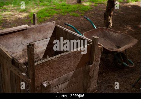 Gros plan du bac à compost en bois près de la brouette vide dans la cour de la maison de campagne. Concept de compostage et d'éco-agriculture Banque D'Images