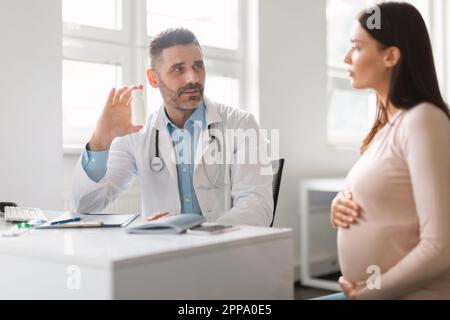 Un médecin d'âge moyen de sexe masculin prescrivant des pilules ou des vitamines pour une femme enceinte lors d'un rendez-vous dans un bureau de clinique moderne Banque D'Images