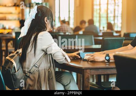 Vue arrière de la femme asiatique avec sac à dos tenant la file d'attente sans fil de la machine à appelants à la main pour attendre la nourriture dans le restaurant. Le pager d'avertissement de signal d'alarme se trouve en mode autonome Banque D'Images