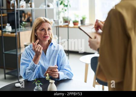 Femme blonde mûre en chemise bleue dans les écouteurs à l'aide d'un smartphone et parlant à une jeune serveuse écrivant les commandes des invités dans le bloc-notes Banque D'Images