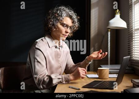 Psychothérapeute mature et confiant parlant au patient lors d'un rendez-vous ou d'une session en ligne, assis devant un ordinateur portable au bureau Banque D'Images