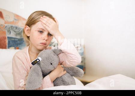 Mesure de la température de l'enfant à l'aide d'un thermomètre numérique. Triste petite fille malade et a mal de tête. Un enfant assis dans un lit qui embrasse un jouet en fourrure, indique un thermomètre Banque D'Images