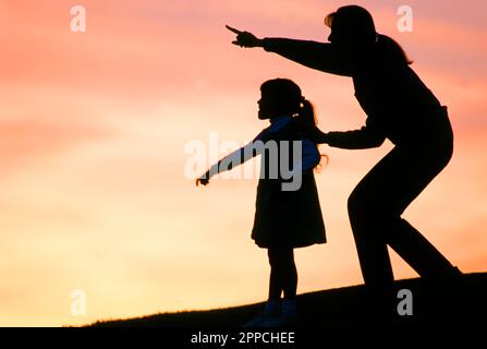 Silhouette d'une mère et d'une fille sur une colline au coucher du soleil avec la mère pointant vers un objet Banque D'Images