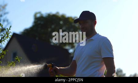 Portrait d'un jeune garçon heureux jardinier tenant dans le tuyau à la main et arrosoir plante verte dans le jardin. Joyeux homme vaporisant de l'eau sur l'arbre par beau temps. Accueil Banque D'Images