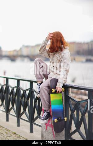 une jeune fille touche ses cheveux rouges assis sur le trottoir et tient son skateboard Banque D'Images