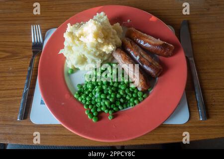 Saucisses, purée de pommes de terre et petits pois sur une assiette rouge, nourriture de confort pour le dîner. Banque D'Images