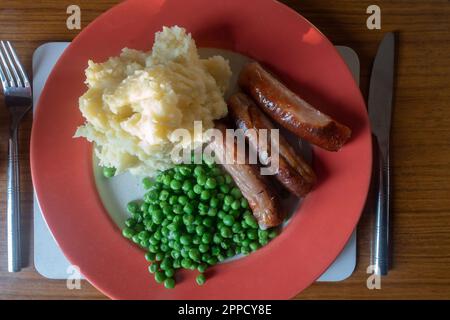 Saucisses, purée de pommes de terre et petits pois sur une assiette rouge, nourriture de confort pour le dîner. Banque D'Images