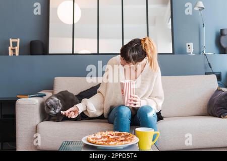 Femme nourrissant du pop-corn pour chat assis sur un canapé à la maison Banque D'Images