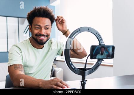 Homme souriant avec la main dans les cheveux de faire tutoriel à travers le smartphone à la maison Banque D'Images