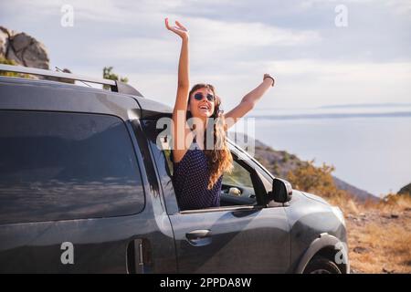 Jeune femme gaie avec les bras relevés et penchée hors de la fenêtre de la voiture Banque D'Images