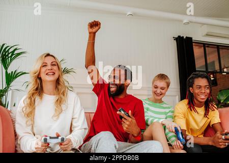 Jeune homme excité avec la main levée assis par des amis jouant à un jeu vidéo à la maison Banque D'Images