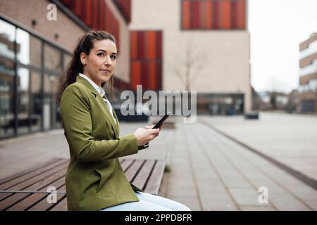 Femme d'affaires attentionnés assise avec un téléphone portable sur un banc à l'extérieur du bâtiment Banque D'Images