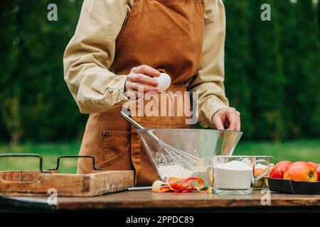 Femme en tablier cassant l'œuf dans le bol à mélanger à la table dans le jardin Banque D'Images