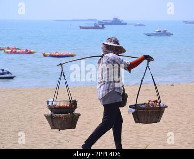 Une femme porte deux paniers sur un poteau sur son épaule pour vendre de la nourriture sur la plage; mer, bateaux, en arrière-plan, à Pattaya, Thaïlande. Travail à chaud quand 33C. Banque D'Images
