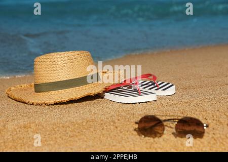 Chapeau de paille élégant, lunettes de soleil et tongs rayées sur la plage de sable près de la mer Banque D'Images